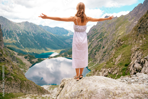 Beautiful long haired girl in white dress standing with hands apart in Polish mountains with fabulous scenic view on background