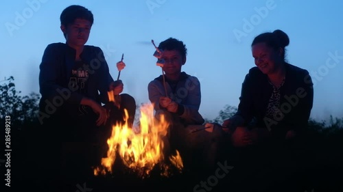 Caucasian mother and two sons sit on camp chairs by the flaming hot fire and fry on a long thin wooden twig square sweet marshmallows. Talking and laughing. Eat hot sweets against the evening sky