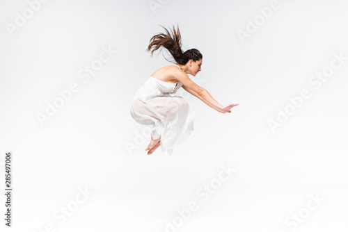 side view of young beautiful ballerina jumping while dancing on grey background