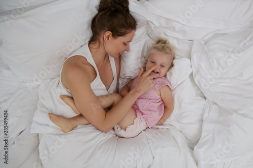 Mother and her infant baby cuddling in the bed, adorable blonde baby and her mum having fun, happy family life concept