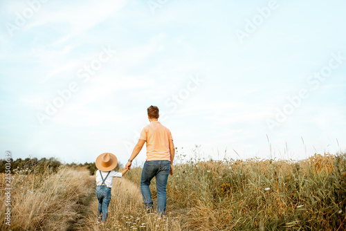 Father and young son walking keeping hands together on the field during the summer activity  back view