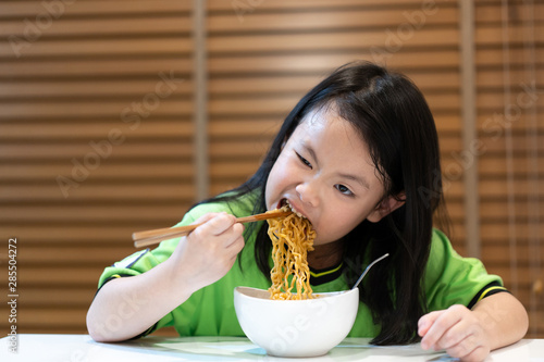 Asian little cute girl eating delicious instant noodles with chopstick by herself on wooden curtain background.Food,Family,Childhood Concept.Instant noodles is a convenient,delicious and cheap food.