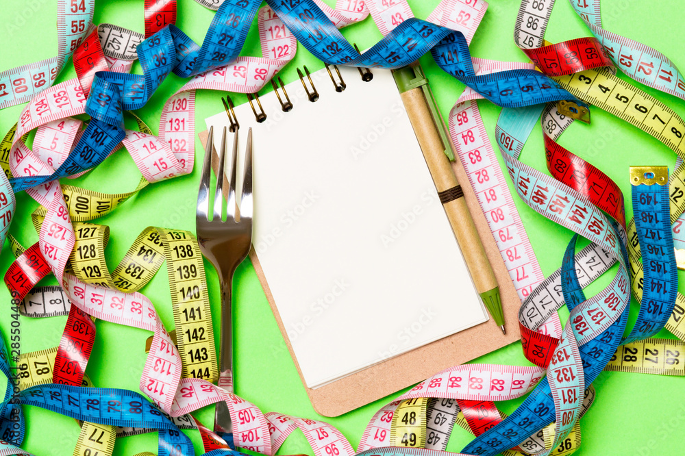 Group of colorful measure tapes, pen, open notebook and fork on green background with empty space for your idea. Top view of healthy lifestyle concept Group of colorful measure tapes, pen, open notebook and fork on green background with empty space for your idea. Top view of healthy lifestyle concept