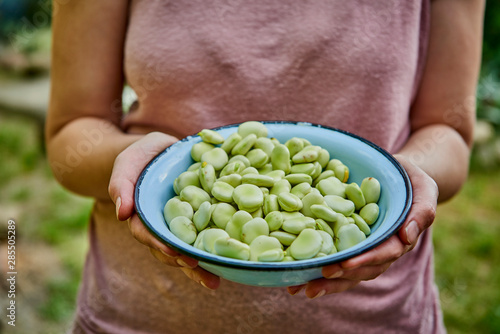 A woman in a pink blouse holds broad beans in a blue bowl