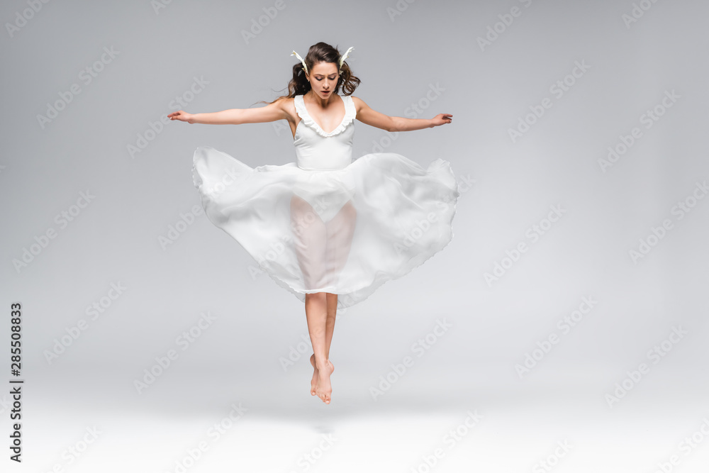 young attractive ballerina in white dress jumping while dancing on grey background