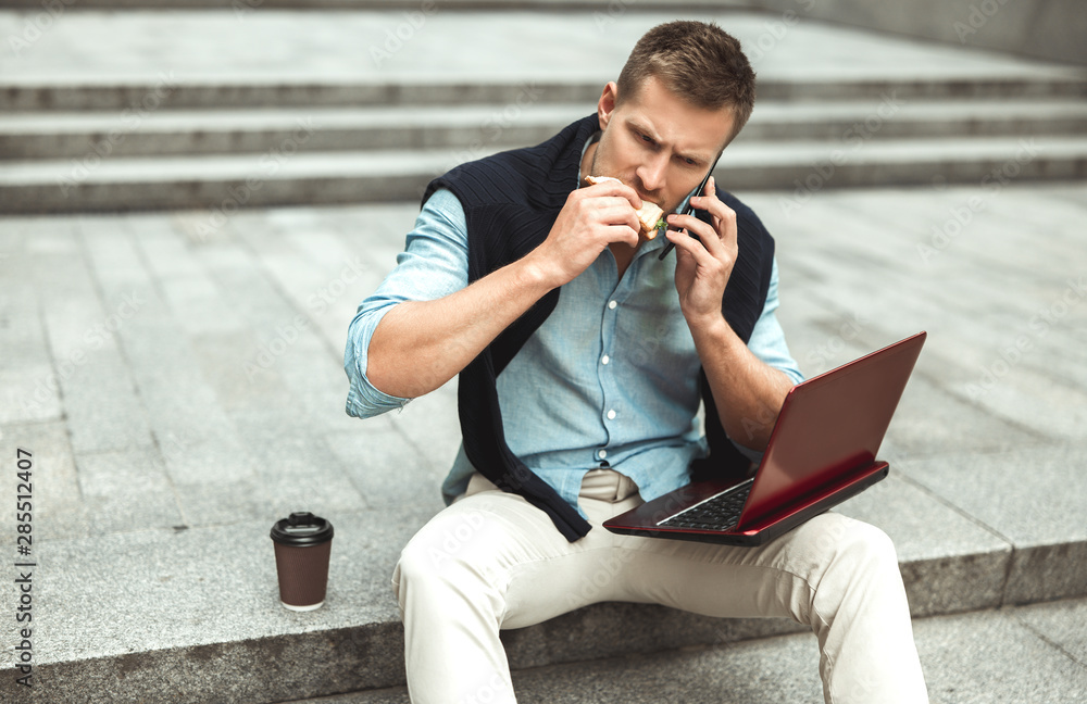 Obraz premium young handsome office worker having conversation while biting his sandwich and drinking coffee for lunch during break sitting on the stairs outside with laptop