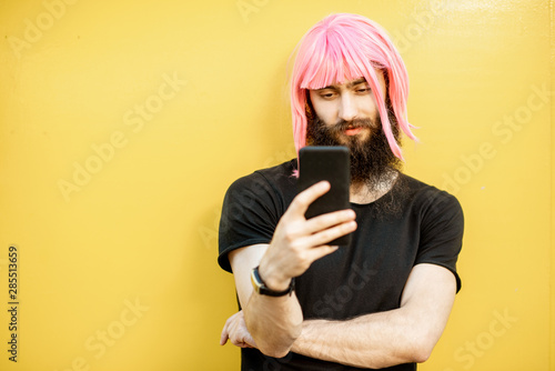 Funny portrait of a stylish man with beard and long color hair usning smart phone on the yellow background photo