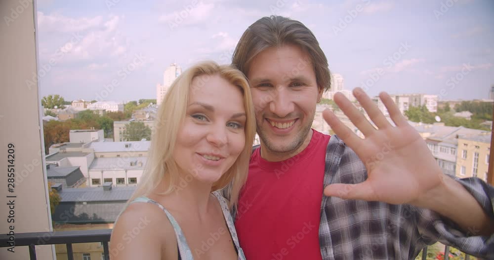 Closeup portrait of happy attractive caucasian couple having video call on phone waving hi hugging standing on balcony of house Closeup portrait of happy attractive caucasian couple having video call on phone waving hi hugging standing on balcony of house