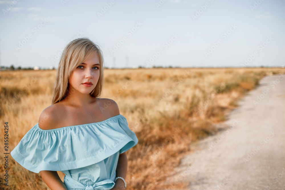 portrait of a young beautiful caucasian blonde girl in a light blue dress standing on a field with sun-dried grass next to a small country road portrait of a young beautiful caucasian blonde girl in a light blue dress standing on a field with sun-dried grass next to a small country road