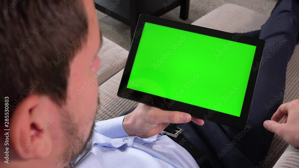 A man sits on a couch in an apartment and works on a tablet with a