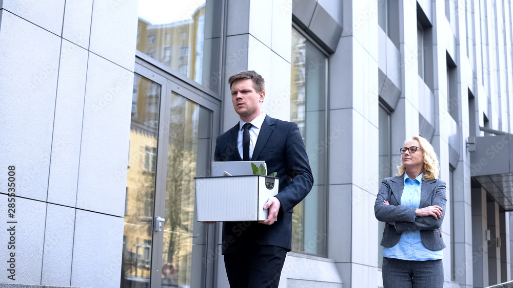 Gloating mature woman looking at dismissed young worker leaving office, conflict Gloating mature woman looking at dismissed young worker leaving office, conflict