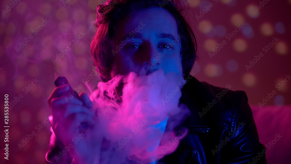 Thoughtful unhappy man exhaling smoke of e-cigarette in nightclub lights, vaping