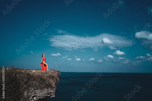Aerial drone view of beautiful woman in red dress standing on the sea shore cliff during sunny summer day