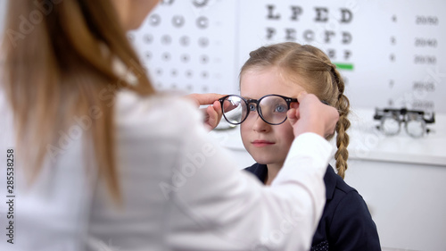 Optician putting glasses on schoolgirl, checking vision and recommending glasses photo