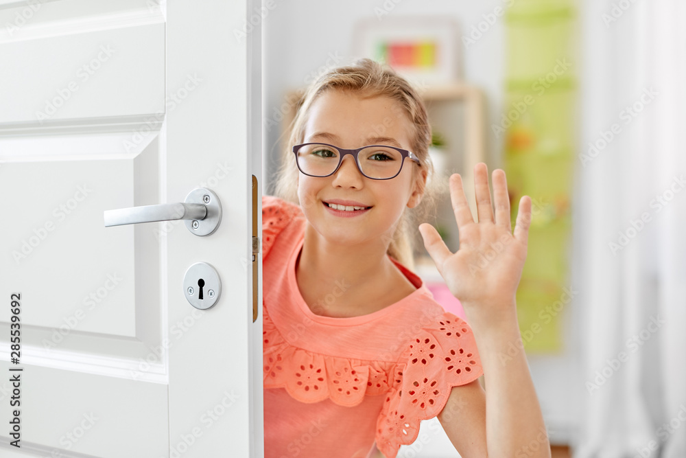 childhood, fun and people concept - happy smiling beautiful girl in glasses behind door waving hand at home
