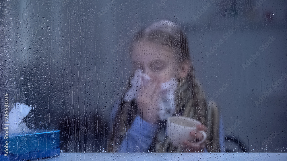 Little girl suffering from flu behind rainy window sneezing and holding tea cup