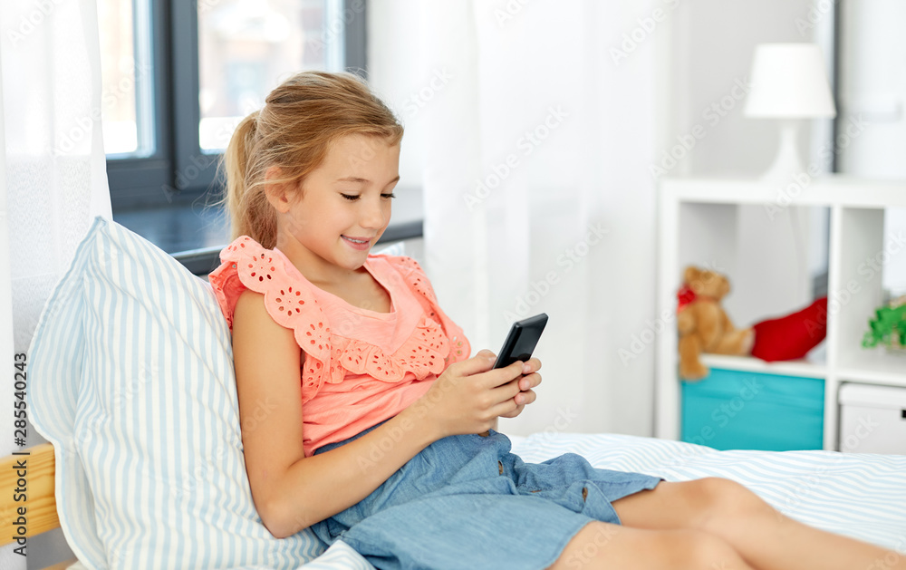 children, technology and communication concept - smiling girl using smartphone sitting on bed at home children, technology and communication concept - smiling girl using smartphone sitting on bed at home