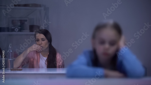 Sad little girl sitting on rainy day, addicted mother drinking alcohol in room