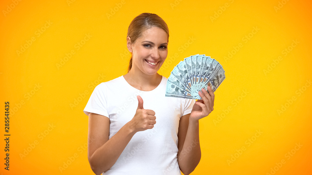 Cheerful woman showing thumbs up holding dollars in hand, well paid job, profit Cheerful woman showing thumbs up holding dollars in hand, well paid job, profit