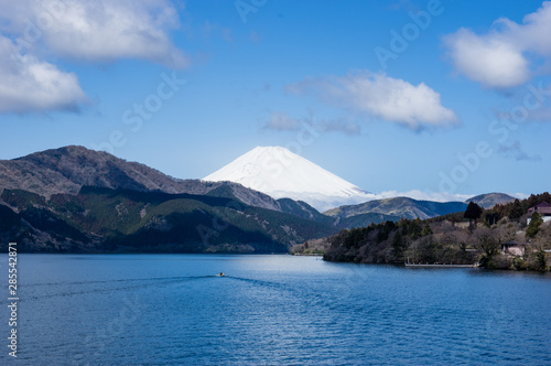 Mount Fuji in Japan covered by snow seen across a large lake. Small boat leaving a triangle form wake as if mirroring the shape of the mountain. Blue sky, white clouds, spring in coastal forests