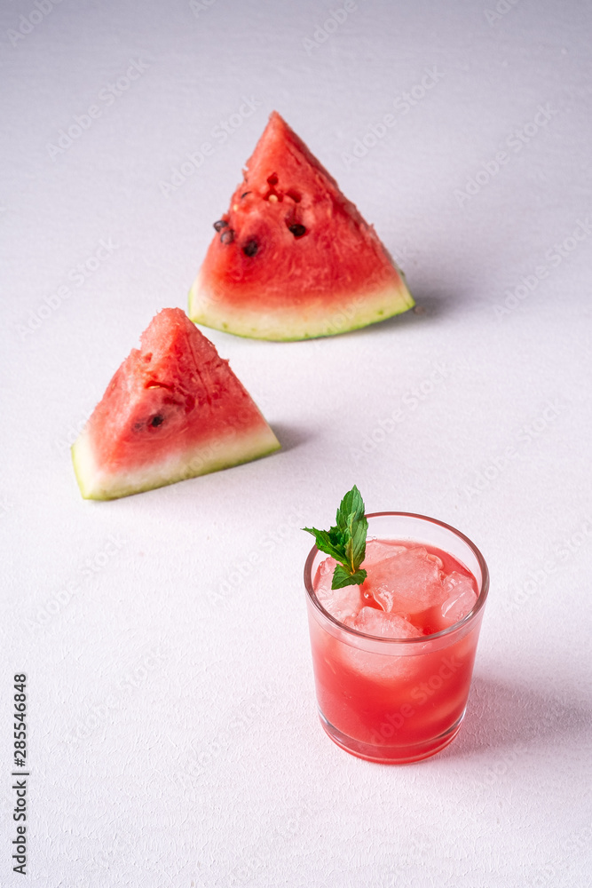 Fresh cold watermelon juice with ice cubes and green mint leaf in glass drink close to two slices of watermelon on white background, copy space, angle view, selective focus Fresh cold watermelon juice with ice cubes and green mint leaf in glass drink close to two slices of watermelon on white background, copy space, angle view, selective focus