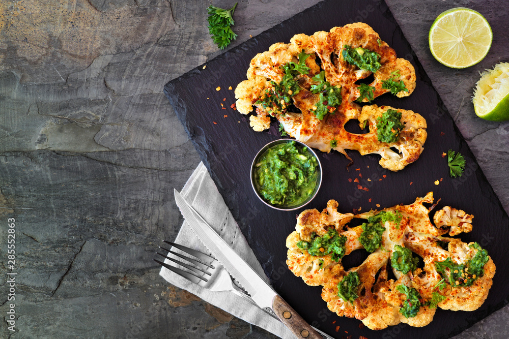 Cauliflower steaks with a cilantro lime sauce. Top view scene on a dark stone background. Healthy eating, plant based meat substitute concept. - obrazy, fototapety, plakaty 