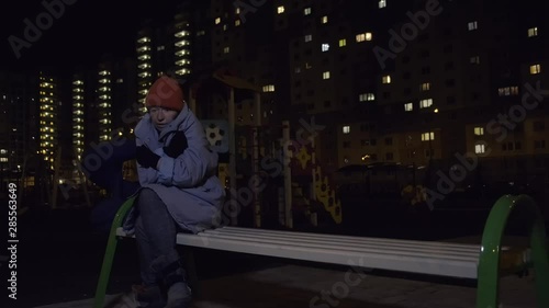 homeless person sits on bench in playground against background of night city. houseless woman is cold and lonely.