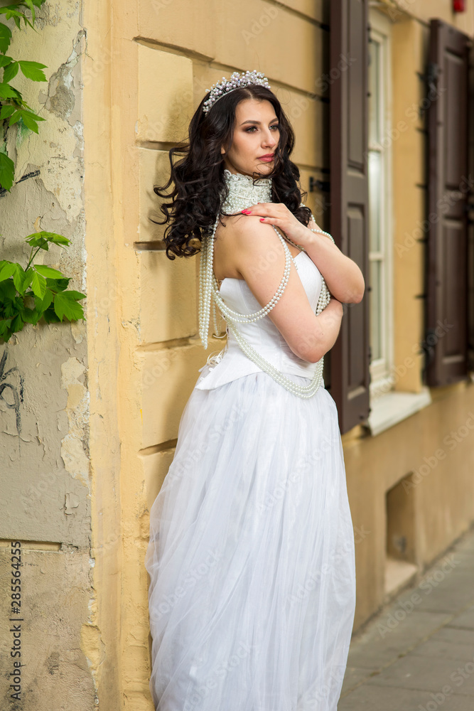 Portrait of Tranquil Caucasian Brunette Bride with Tiara Posing Against Old Building. Wearing Beads Necklace and Decorations. Portrait of Tranquil Caucasian Brunette Bride with Tiara Posing Against Old Building. Wearing Beads Necklace and Decorations.