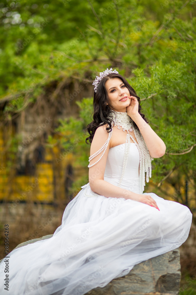 Portrait of Sensual Caucasian Bride With Diadem Sitting On Stone In Flowers Garden Outdoors. Wearing Beads Necklace. Portrait of Sensual Caucasian Bride With Diadem Sitting On Stone In Flowers Garden Outdoors. Wearing Beads Necklace.