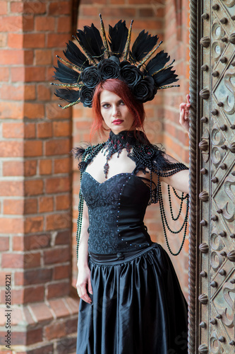 Tranquil Gothic Girl in Long Black Dress. Wearing Artistic Feather Crown with Roses. Posing Against Old Castle Gates.
