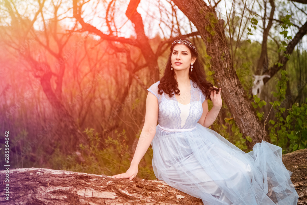 Gorgeous Portrait of Sensual Caucasian Brunette in Long Blue Dress Posing on Wood Pile Against Bright Light.