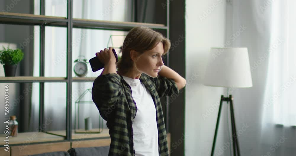 Young woman is putting wireless headset on, using gadget to switch on mp3 application, going to listen music, smiling, standing in living room, indoor. Caucasian, 4K, shot on RED camera. Young woman is putting wireless headset on, using gadget to switch on mp3 application, going to listen music, smiling, standing in living room, indoor. Caucasian, 4K, shot on RED camera.