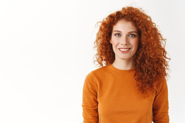 Happy cute redhead woman with curly hair smiling sincere and carefree, standing white background wear orange sweater, look camera normal casual attitude, customer support answer questions politely