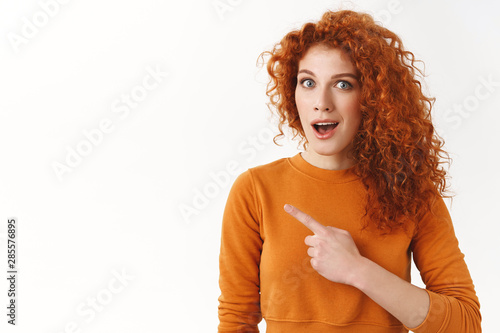 Excited curious redhead curly-haired gorgeous woman looking questioned, open mouth gasping intrigued, pointing finger left astonished, discuss breathtaking news, stand white background