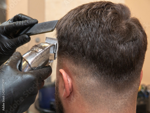 Hairstylist working with clipper and comb, close up view. Mans head and masters hands in black rubber gloves. Stylist at work in studio. Selective soft focus. Blurred background