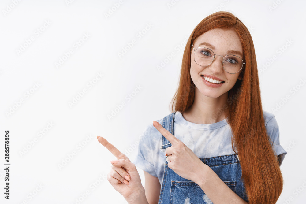 Obraz premium Waist-up silly cheerful redhead female asking question tilt head intrigued, stand white background, pointing left, showing promo, questioned discuss interesting advertisement white background