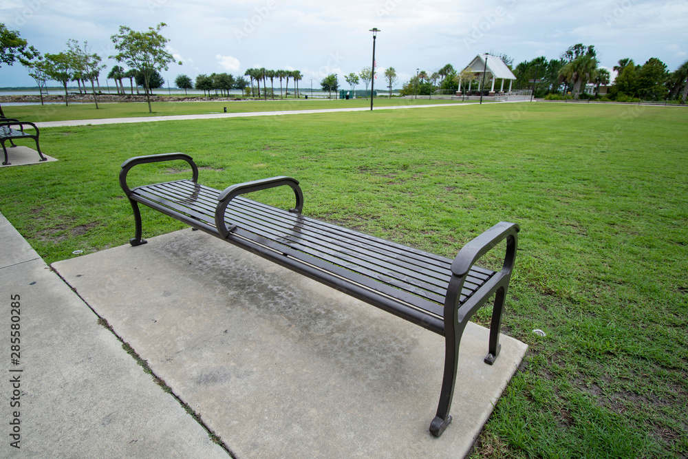 A close up perspective of a dark metal park bench meant to sit on surrounded by the concrete sidewalk surrounded by bright green grass outside on a cloudy day with a pavilion in the background.