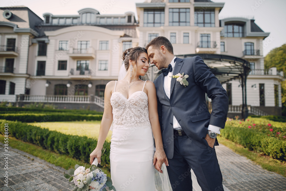 A young and beautiful bride and her husband is standing in a summer park with bouquet of flowers