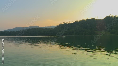 Wide low-angle panoramic still shot from Temenngor lake, of partial silhouettes of forest covered landscape, and a boat sailing on the serene waters of the Lake, Garik,  Malaysia photo