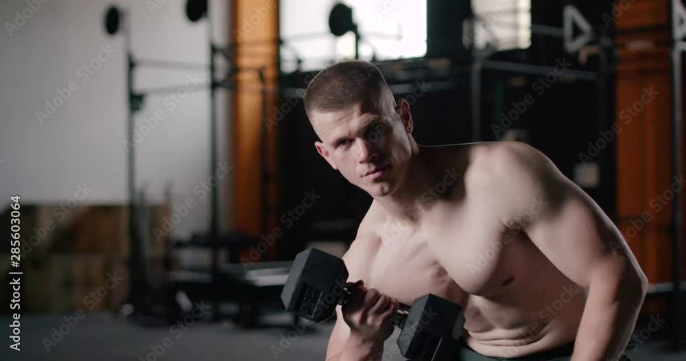 Muscular man in Gym lifts the dumbbells, man using dumbbells for biceps exercises and looks into the camera. Muscular man in Gym lifts the dumbbells, man using dumbbells for biceps exercises and looks into the camera.