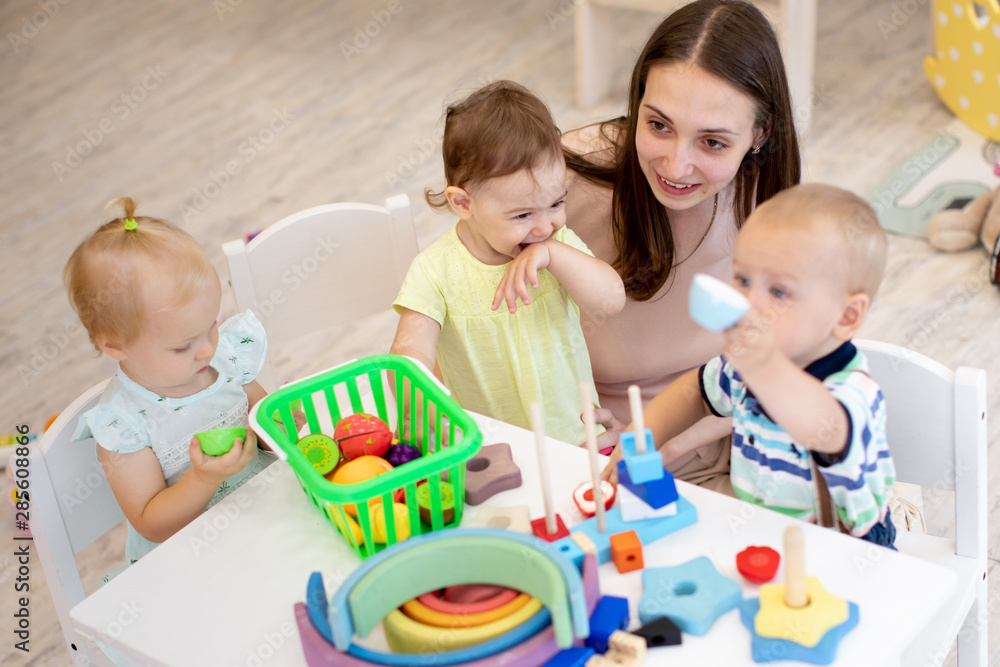 Fototapeta premium Nursery babies playing with teacher in kindergarten. Top view of kids with educator in creche