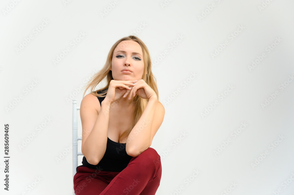 Studio waist-length photo portrait of a pretty beautiful young happy blonde woman with long hair on a white background in a black t-shirt and red trousers. Sits on a chair, smiles, shows emotions. Studio waist-length photo portrait of a pretty beautiful young happy blonde woman with long hair on a white background in a black t-shirt and red trousers. Sits on a chair, smiles, shows emotions.