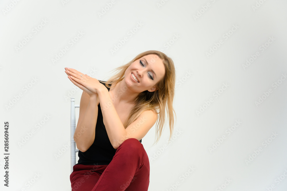 Studio waist-length photo portrait of a pretty beautiful young happy blonde woman with long hair on a white background in a black t-shirt and red trousers. Sits on a chair, smiles, shows emotions. Studio waist-length photo portrait of a pretty beautiful young happy blonde woman with long hair on a white background in a black t-shirt and red trousers. Sits on a chair, smiles, shows emotions.