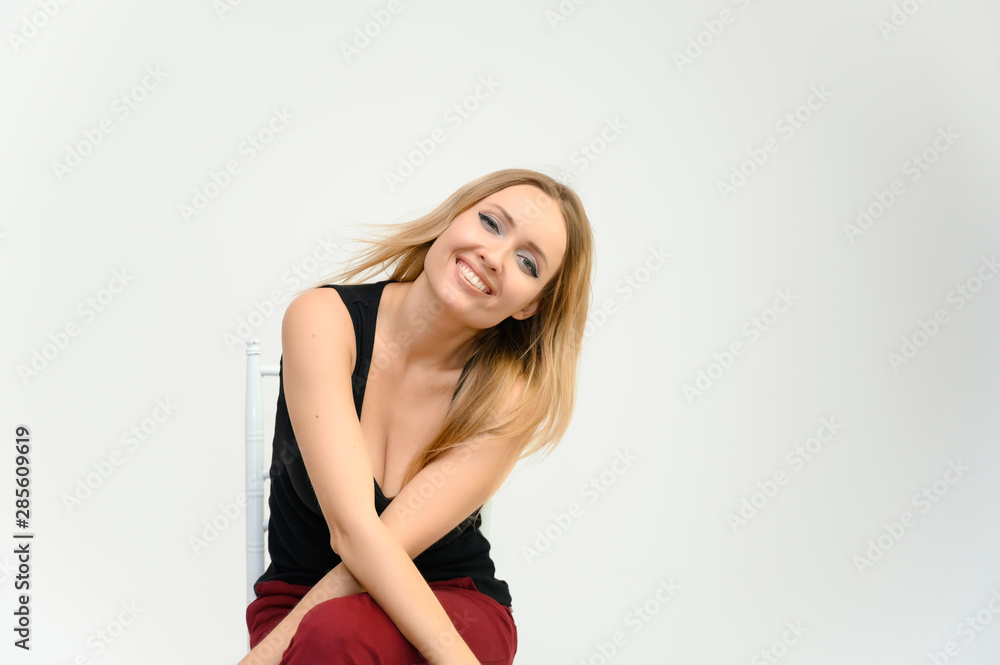 Studio waist-length photo portrait of a pretty beautiful young happy blonde woman with long hair on a white background in a black t-shirt and red trousers. Sits on a chair, smiles, shows emotions. Studio waist-length photo portrait of a pretty beautiful young happy blonde woman with long hair on a white background in a black t-shirt and red trousers. Sits on a chair, smiles, shows emotions.