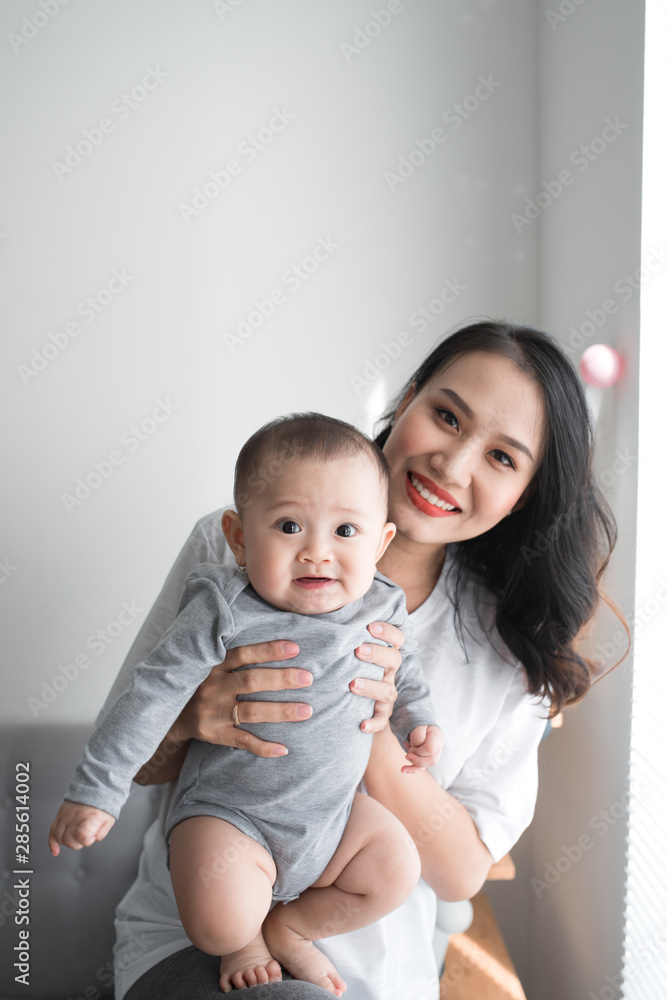 Beautiful young mom is playing with her cute baby and smiling while standing near the window at home Beautiful young mom is playing with her cute baby and smiling while standing near the window at home