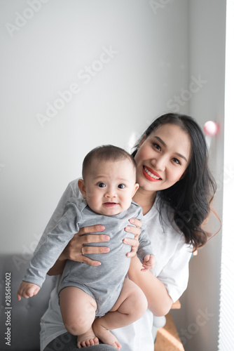 Beautiful young mom is playing with her cute baby and smiling while standing near the window at home