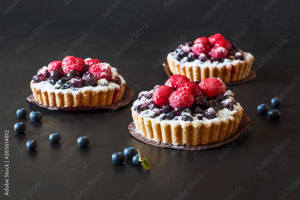 Three delicious tarts, colorful pastry cakes sweets with fresh raspberries and blueberry on black background.