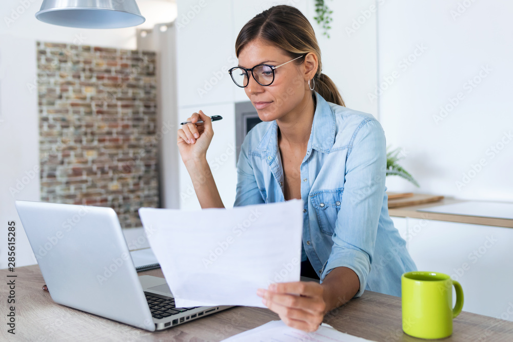 Pretty young woman working with laptop and documents in the kitchen at home. Pretty young woman working with laptop and documents in the kitchen at home.