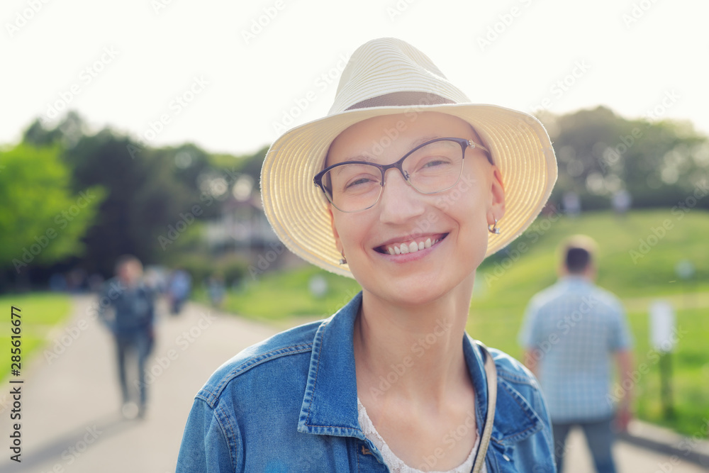 Happy young caucasian bald woman in hat and casual clothes enjoying life  after surviving breast cancer. Portrait of beautiful hairless girl smiling  during walk at city park after curing disease Stock Photo |