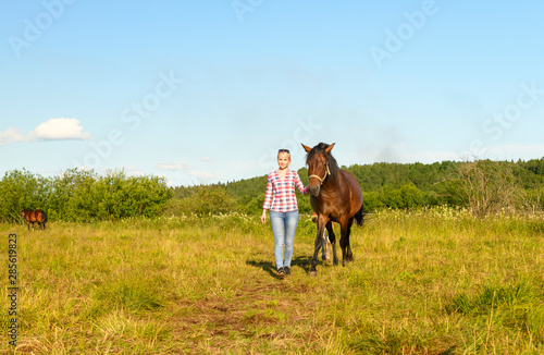 The smiling Caucasian woman is holding her bay horse by the natural halter and leading from a pasture.